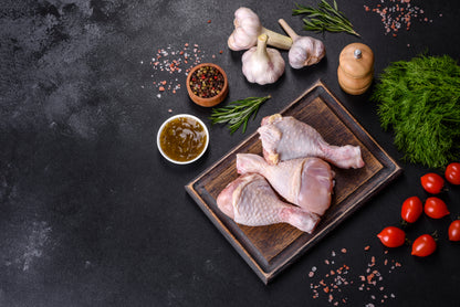 Raw chicken legs on a wooden board with herbs, spices, and tomatoes on a dark surface